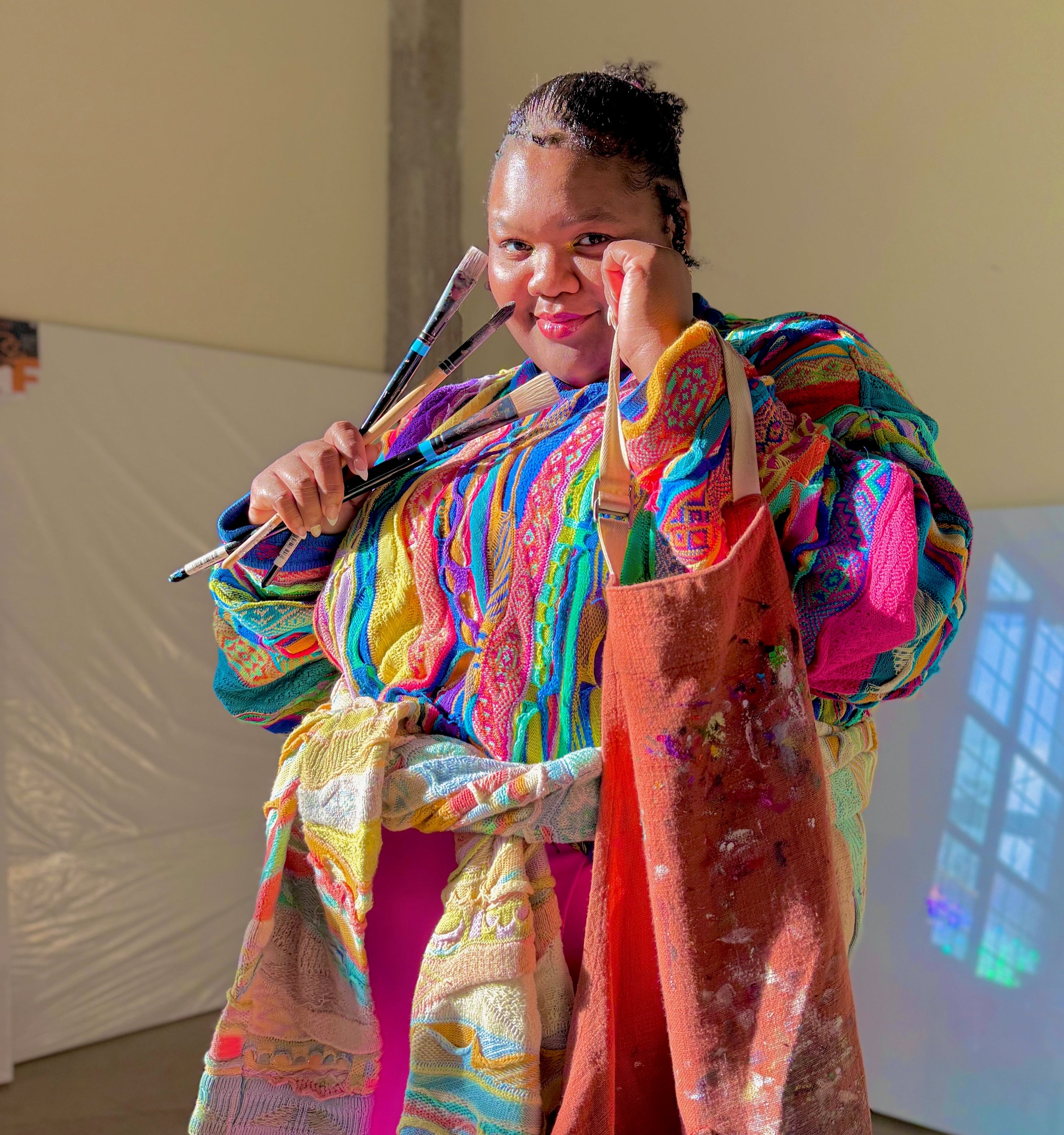 LaNia the Artist wearing a colorful outfit with an apron, holding paintbrushes in front of two large canvases.